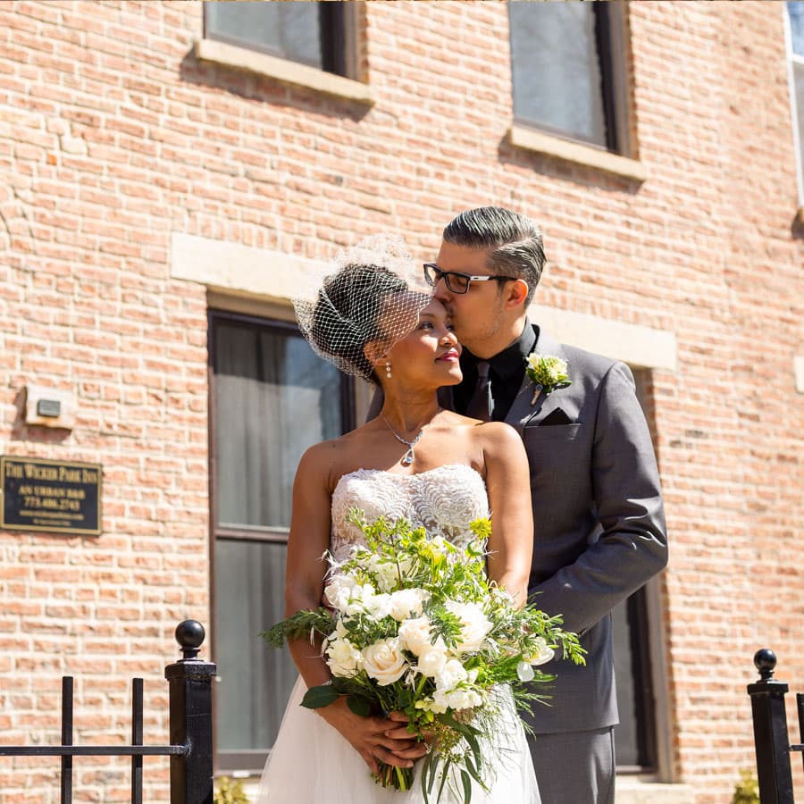 A bride and groom embrace lovingly outside a brick building, with the bride holding a bouquet of white flowers.