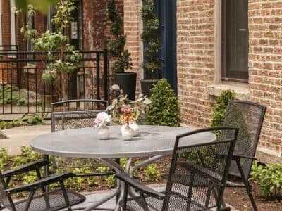 A round metal table with four chairs, adorned with a small flower arrangement, set against a brick wall backdrop.
