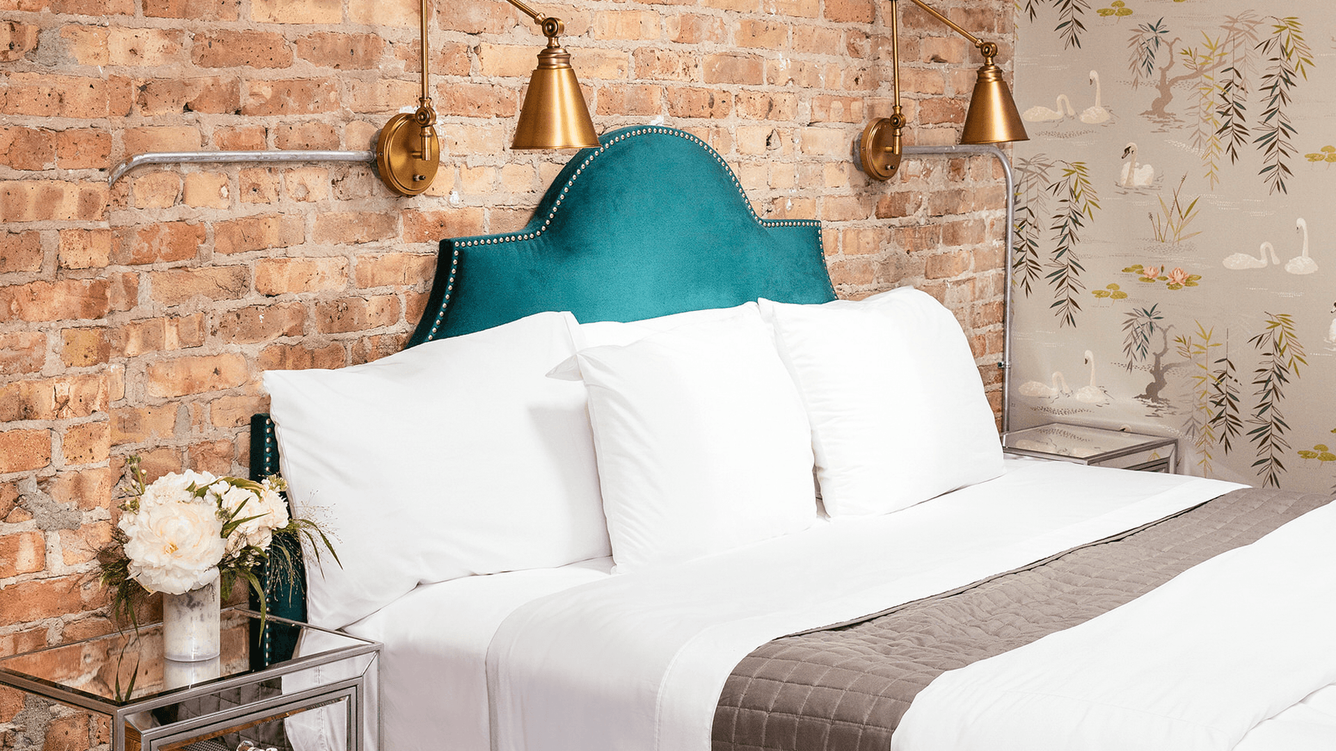 Elegant bedroom featuring a teal headboard, white bedding, stylish lamps, and a floral arrangement against a brick wall.