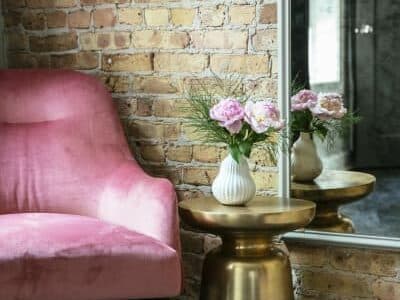 A pink velvet chair beside a gold accent table with a vase of flowers, in front of a brick wall and mirror.