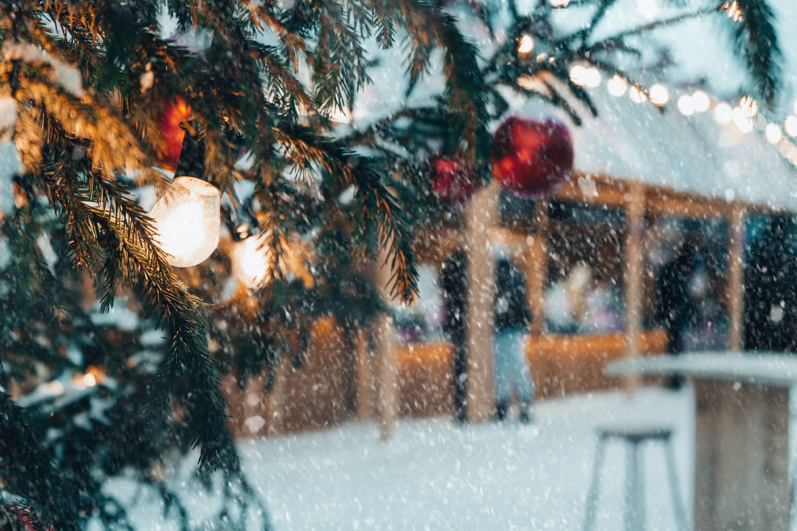 A snowy scene featuring a decorated evergreen tree with lights and ornaments, set against a blurred market backdrop.