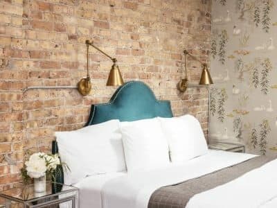 A stylish bedroom featuring a tufted teal headboard, white bedding, and exposed brick walls with gold wall sconces.